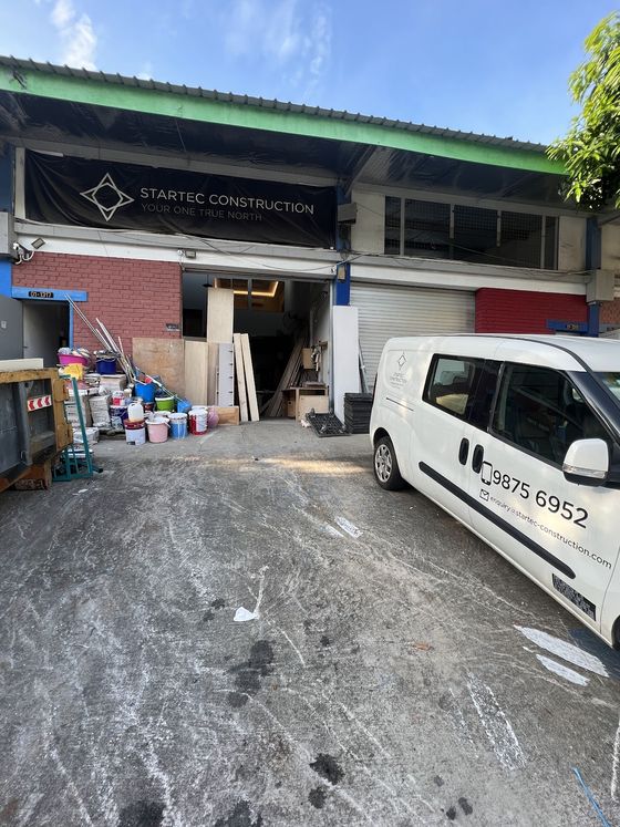 Startec Construction's own factory unit at Toa Payoh Industrial Park: a black banner over the roller shutter, plywood and timber visible inside, and the Startec van parked outside.